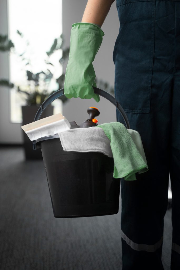 A person in dark blue overalls and green gloves holds a black bucket filled with cleaning supplies.
