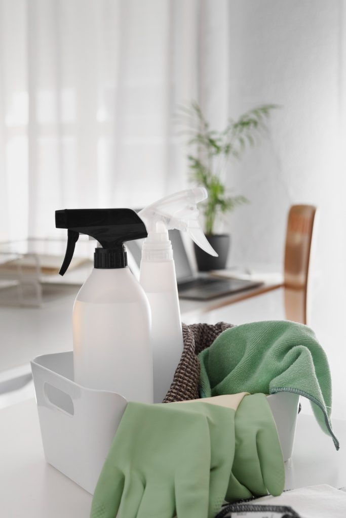 A white caddy holds cleaning supplies: two spray bottles, green and brown cloths, and green gloves.