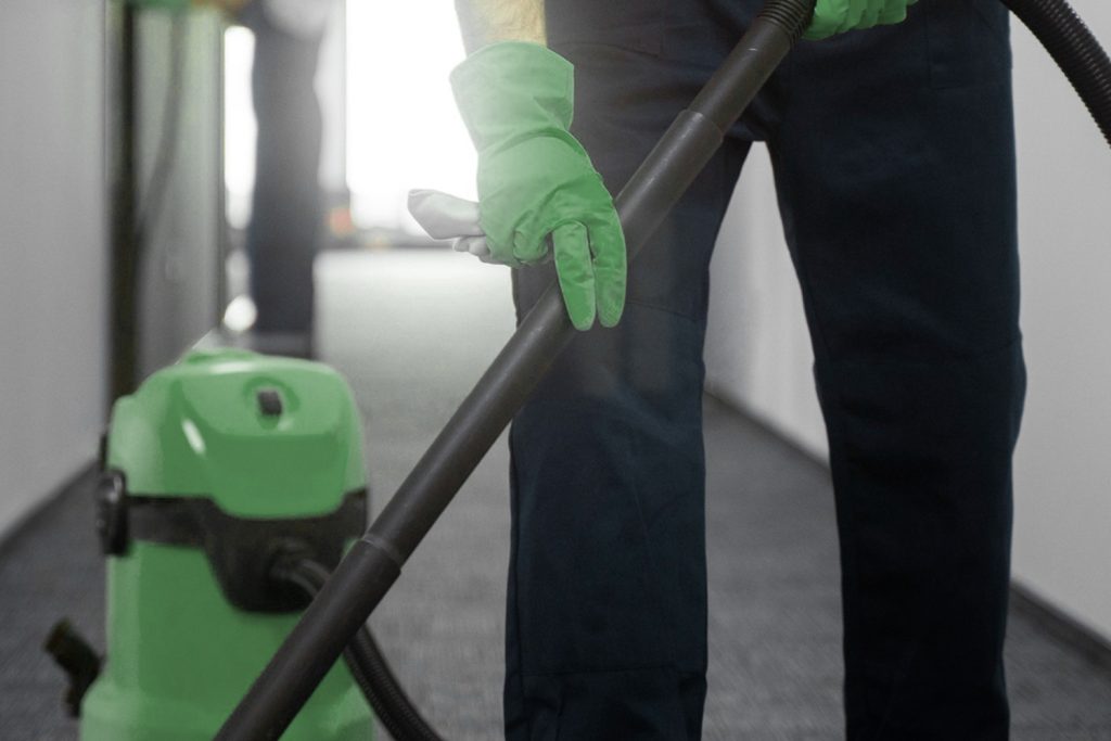 A person in green gloves operates a green and black wet/dry vacuum cleaner on a tiled floor.